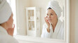 © Taha - Portrait of mature woman examining face skin in a mirror, wearing a white robe and towel in bright room. Reflection in a mirror of a beautiful woman applying cosmetic skin care