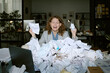 © AnnaStills - Caucasian young woman sitting at cluttered desk holding crumpled papers, appearing stressed while surrounded by large pile of documents in modern business office environment