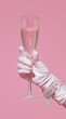 © sornram - A stylish hand in a white glove holding a champagne glass against a soft pink background, exuding elegance and celebration.