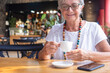© luciano - Attractive senior woman relaxing for a break at coffee shop in mall holding a coffee cup. Carefree smiling elderly woman with eyeglasses and necklace