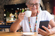 © luciano - Attractive senior woman relaxing for a break at coffee shop in mall using mobile phone holding a coffee cup. Carefree smiling elderly woman with eyeglasses and necklace