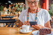 © luciano - Smiling senior woman relaxing for a break at coffee shop with chocolate cake and coffee cup. Carefree elderly woman with eyeglasses and necklace using mobile phone