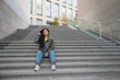 © Татьяна Зернова - Young Asian woman with long black hair sits on stone steps in an urban setting. She wears a leather jacket, yellow shirt, and blue jeans, with white sneakers.