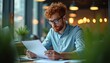 © Viktor - Man with red hair and glasses reads documents at desk. Laptop and plants nearby. Modern office interior with warm lights. Focused professional works on paper.