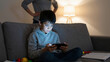 © Lubov - Young boy immersed in smartphone screen time on a sofa. A concerned parent observes, highlighting digital addiction and technology impact on family life