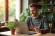 © Viktor - Young man happily shops online using laptop and credit card. He sits at table with houseplants near window. Natural light illuminates comfortable home workspace and happy shopper.