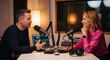© Khan - Two people are sitting at a table in a recording studio. A man is speaking into a microphone while a woman listens attentively, smiling.