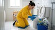 © Coosh448 - A woman in a yellow protective suit and blue rubber gloves kneeling beside a white ceramic toilet bowl, scrubbing the interior with a blue microfiber cloth, cleaning spray and supply bucket visible on