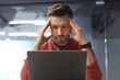 © Prostock-studio - A man is sitting at a desk with a laptop in front of him. He holds his head with his hands and looks concerned. The setting is a modern office space with glass walls.