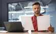 © Prostock-studio - A man sits at a desk with a laptop and holds two documents. He appears focused as he looks closely at the papers. The setting is a modern office with bright lights.