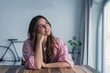 © Daniel - A young woman in a pink shirt sits at a rustic wooden table by a large window, smiling as she daydreams and reflects in a bright, cozy apartment with a bicycle and houseplant in the background