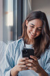 © Daniel - A young woman sits by a window smiling as she scrolls and texts on her smartphone, conveying relaxation, connection and modern lifestyle in a bright indoor setting.