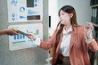 © MINAE - Young businesswoman with a pen in hand receiving a smartphone from a colleague, standing in an office with data charts pinned on a grey board, showing business analytics and financial reports