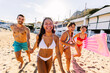 © Xavier Lorenzo - Diverse group of friends laughing and running along a sunny sandy beach in swimwear, enjoying carefree summer vacation together with mountains and resort buildings in the background
