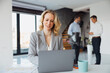© Novak - Blonde businesswoman focused on her laptop at a desk while two male colleagues stand and talk in the background.