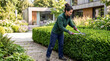 © Artem Zatsepilin - caucasian woman in green jacket and blue pants trimming hedge with shears in backyard. gardening and landscaping concept. home maintenance, outdoor work. banner, website header, copyspace