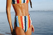 © SHOTPRIME STUDIO - A woman in a colorful striped bikini stands by the water, her skin glistening with moisture. The lifestyle image highlights summer, beach, and healthy skin concepts for women.