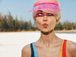 © SHOTPRIME STUDIO - A young woman stands on a sunny beach, wearing a pink visor and colorful swimsuit. She blows a kiss toward the camera, radiating playful confidence and warm energy.