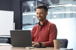 © Prostock-studio - A man sits at a table in an office working on a laptop. He smiles while typing and has a coffee cup beside him. The setting is bright and modern with large windows.
