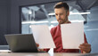 © Prostock-studio - A man sits at a desk in an office looking at two sheets of paper. He appears focused while comparing the documents with a laptop in front of him. Sunlight enters the office space.