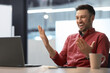 © Prostock-studio - A man wearing a red shirt sits in front of a laptop in an office. He smiles and gestures while having a video call. A coffee cup sits on the table.