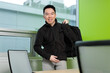© fotolite - A young Asian male student smiles at the camera while putting backpack on in a campus study room, ideal for back to school, student lifestyle, or campus life concepts.