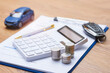 © vadish - Car insurance document on clipboard with white calculator, stacked coins, blue toy car and modern car key on wooden desk. Concept auto insurance premium calculation and vehicle finance planning.