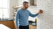 © Balqis - Bearded man in blue sweater adjusting smart thermostat in a modern kitchen
