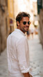 © Olha Havelia - handsome men walking down a narrow European street in a linen shirt and sunglasses, cobblestone floor, vintage warm tones, candid photo while turning back with a subtle smile