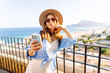 © maxbelchenko - Portrait of a young tourist in a hat holding a phone on a viewing platform. A beautiful woman enjoys the sunset with a smartphone outdoors. Concept: tourism, recreation, and lifestyle.