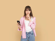 © Designerant - Caucasian woman holding smartphone and making silence gesture with finger on lips, asking for quiet or secrecy concept, wearing pink shirt and denim, isolated on beige background with copy space.