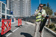 © qunica.com - A construction worker wearing a high-visibility vest talks on a phone while holding a survey tube. He stands at an urban work site near red barriers and modern apartment buildings.