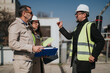 © qunica.com - Three construction professionals wearing hard hats and high-visibility vests discuss project documents on site. They review plans and coordinate work during an outdoor site meeting.