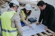 © qunica.com - Construction engineers in hard hats review a large blueprint on a wooden pallet at a work site. The team discusses plans and layout details during an on-site planning meeting.