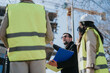 © qunica.com - Construction engineers in high-visibility vests and hard hats review plans at a building site. A project supervisor gestures while colleagues listen during an on-site planning discussion.
