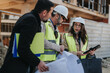 © qunica.com - Construction engineers in safety vests and hard hats review blueprints and a tablet while discussing plans.