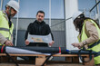 © qunica.com - Construction engineers review a large blueprint while standing at an urban building site. A project manager and site workers discuss plans wearing hard hats and high-visibility vests.