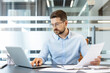 © Liubomir - Businessman wearing eyeglasses and a blue shirt, concentrating on his laptop and carefully reviewing paperwork while sitting at a desk in a modern corporate office environment