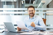 © Liubomir - Happy businessman cheering with a fist pump and a wide smile, holding official document in hand while sitting at a desk with a laptop and paperwork, celebrating good news and career achievement