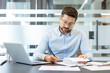 © Liubomir - Businessman in glasses sitting at office desk, reviewing and signing paperwork, working on a laptop with focus and concentration, managing corporate tasks