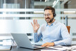 © Liubomir - Man wearing a headset and spectacles, smiling and waving hand to laptop screen, participating in a remote work virtual meeting from his modern office desk