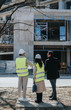 © qunica.com - Two construction engineers in hard hats and high-visibility vests inspect a building under construction. They stand outdoors discussing the project and reviewing plans near the concrete structure.
