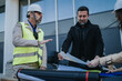 © qunica.com - Two construction engineers discuss and review large architectural plans at a site table. The colleagues examine blueprints together during an outdoor project meeting.