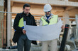 © qunica.com - Two construction engineers review a large blueprint at an outdoor building site. The professionals inspect plans together while wearing safety gear and holding documents.