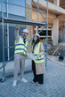 © qunica.com - Construction professionals in hard hats and high-visibility vests review blueprints while standing at a building site. They examine project plans and point toward the structure under construction.