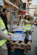 © qunica.com - A construction engineer in a hard hat and hi vis vest reviews blueprints with colleagues at a building site. The team discusses plans and site details around a temporary fence and pallet workspace.