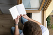 © Diatlova Tetiana - Top view of man reading book by window with natural light in cozy home interior