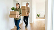 © Taras Roshchuk - A couple walks down a hallway carrying boxes and a small plant while moving into a new home. They smile at each other and look excited about their new space