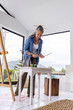 © wavebreak3 - Senior female artist standing at home studio table wearing denim shirt arranging brushes near easel