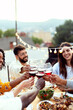 © Xavier Lorenzo - Group of diverse friends enjoying an outdoor summer celebration, clinking red wine glasses in a toast on a rooftop terrace with delicious food and festive string lights